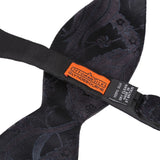 Close-up of a black silk bow tie with a subtle navy blue paisley pattern, featuring an orange and black brand label that reads 'OTTOMAN SILKS GLYDEBOURNE'.